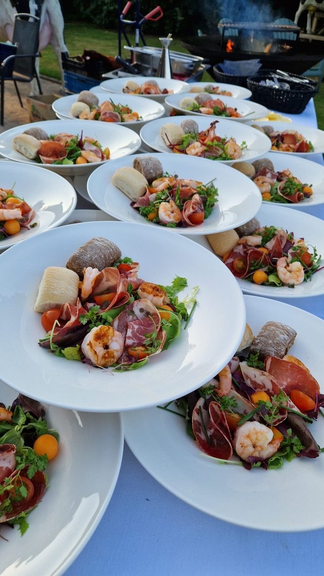 Gedekte tafel buiten met diepe borden salade met garnalen, ham en meloen, klaar om uit te serveren.
