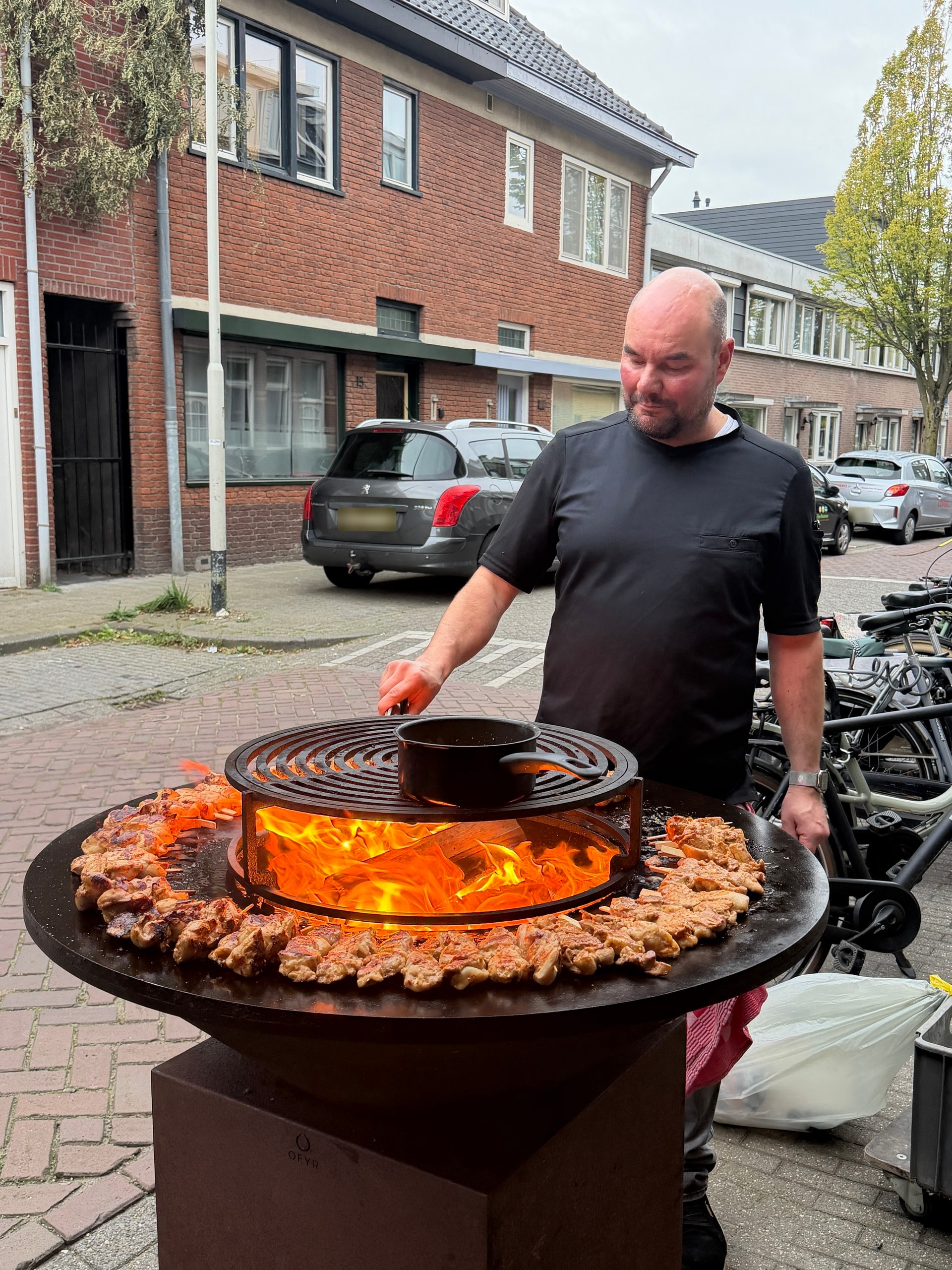 Maarten van Gils kookt buiten op locatie aan een ronde vuurplaat met open vuur en spiesen.
